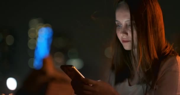 Una chica joven en el fondo de la ciudad nocturna mira en la pantalla del teléfono inteligente — Vídeo de stock