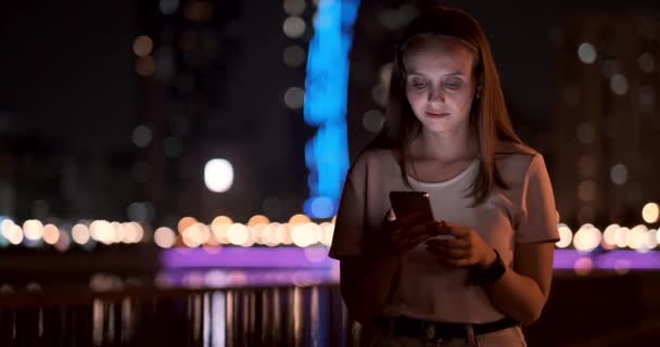 En ung flicka på bakgrunden av Night City tittar in i smartphone skärmen — Stockvideo