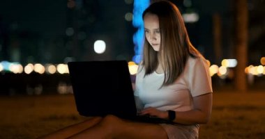 Kadın müteahhit gece şehirde parkta oturur ve dizüstü bilgisayara bakarak kod yazar.