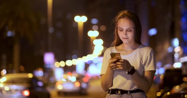 Een jong meisje op de achtergrond van de nacht stad kijkt naar het scherm van de smartphone — Stockvideo