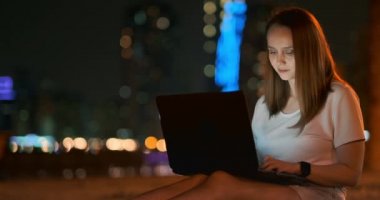 Genç tasarımcı kız geceleri dışarıda, palmiye ağaçlarının altında dizüstü bilgisayarla çalışıyor.