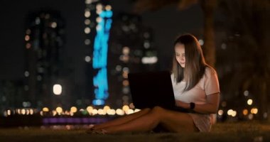 Genç tasarımcı kız geceleri dışarıda, palmiye ağaçlarının altında dizüstü bilgisayarla çalışıyor.