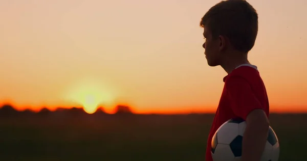 Ung fotbollsspelare går med bollen på planen drömmer om en fotbollskarriär, vid solnedgången tittar på solen — Stockfoto