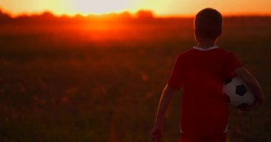 Kamera bir futbol topu ile gün batımında alan boyunca yürüyen çocuk izler.