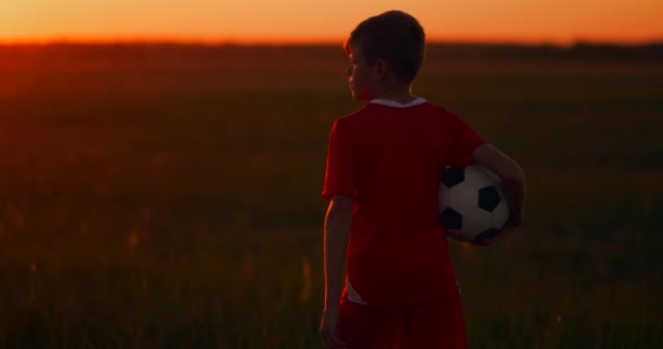 A câmera segue o garoto andando ao pôr do sol no campo com a bola . — Vídeo de Stock