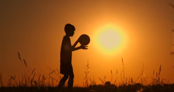 Pequeño jugador de fútbol silueta, niño hace malabares con una pelota en el campo al atardecer. cámara lenta . — Vídeos de Stock