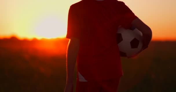 Niño sosteniendo una pelota de fútbol de pie al atardecer en el campo — Vídeo de stock