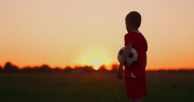 Erkek futbolcu topu sahada, batan güneş ikamerayla çocuğu takip ediyor.