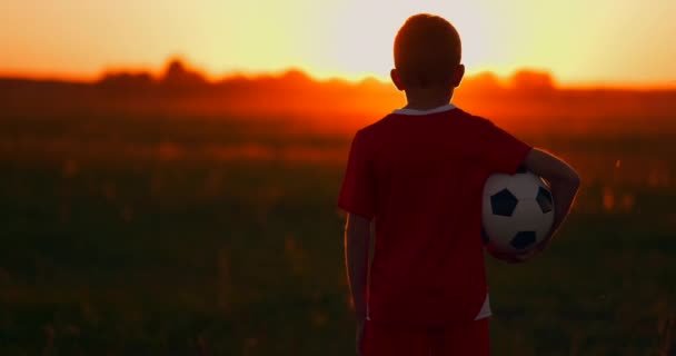 Pojke med en boll i ett fält i solnedgången, pojke drömmar om att bli en fotbollsspelare, går pojken till fältet med bollen vid solnedgången — Stockvideo