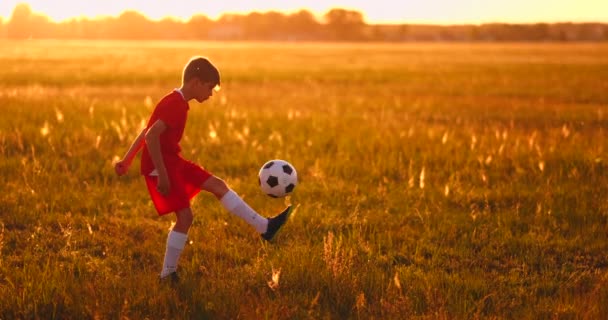 Niño jugador de fútbol al atardecer haciendo malabares con la pelota en el campo — Vídeos de Stock