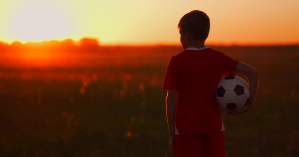 Niño con una pelota en un campo al atardecer, niño sueña con convertirse en un jugador de fútbol, niño va al campo con la pelota al atardecer — Vídeos de Stock