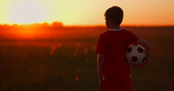 Pojke med en boll i ett fält i solnedgången, pojke drömmar om att bli en fotbollsspelare, går pojken till fältet med bollen vid solnedgången — Stockvideo