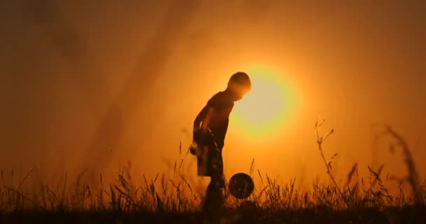 Pequeño jugador de fútbol silueta, niño hace malabares con una pelota en el campo al atardecer. cámara lenta . — Vídeo de stock