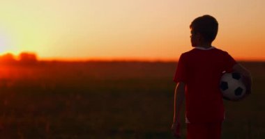Kamera, gün batımında sahada topla yürüyen çocuğu takip ediyor..