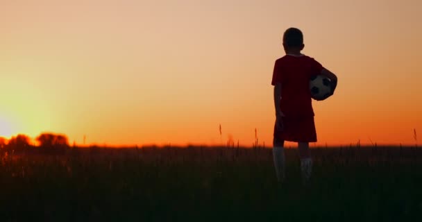 Um menino com uma bola de futebol vai para o campo e olha para o nascer do sol . — Vídeo de Stock