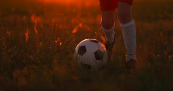 Rastreando um menino jogador de futebol correndo com uma bola na câmera ao pôr do sol — Vídeo de Stock