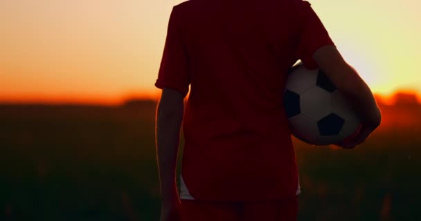 Menino segurando uma bola de futebol de pé ao pôr do sol no campo — Vídeo de Stock