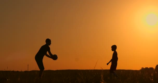 Отец и сын играют в футбол в парке на закате, силуэты на фоне яркого солнца, замедленная съемка — стоковое видео