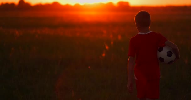 Chłopiec z piłką o zachodzie słońca idzie na słońce i wygląda. — Wideo stockowe