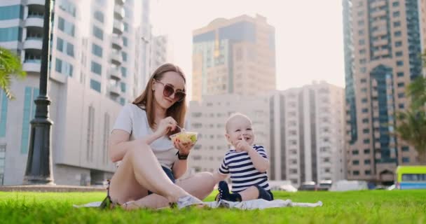 Молодая мама с ребенком, сидящая на траве в парке обедает — стоковое видео