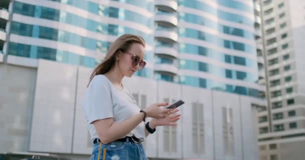 Молодая красивая женщина улыбается со смартфоном в руке против летних небоскребов — стоковое видео