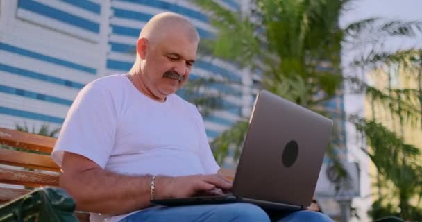 Hombre mayor sentado en un banco del parque entre las palmeras y mirando la pantalla del ordenador portátil — Vídeo de stock