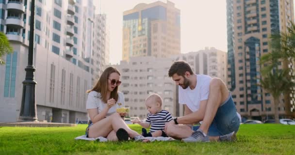 Сім'я сидить на траві в місті, сміючись з маленькою дитиною . — стокове відео