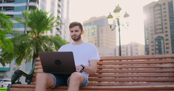 A young man in the summer of palm trees sitting with a laptop and typing on the keyboard — Stock Video