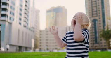 Erkek bebek yaz arka plan binalarda çim üzerinde oturan gülüyor