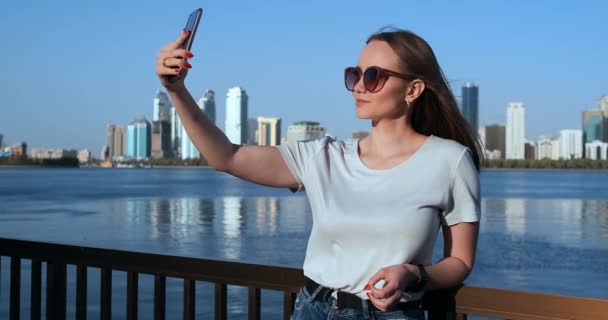 Летом девушка на фоне большого города и набережной делает селфи на смартфоне — стоковое видео
