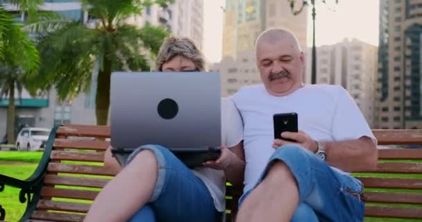 Casal sênior feliz sentado em um banco no verão em uma cidade moderna com um laptop no fundo de arranha-céus — Vídeo de Stock