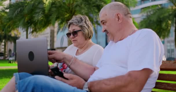 Feliz pareja de ancianos sentados en un banco en el verano en una ciudad moderna con un ordenador portátil en el fondo de los rascacielos — Vídeo de stock