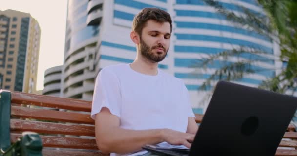 Un joven en el verano de palmeras sentado con un ordenador portátil y escribiendo en el teclado — Vídeo de stock