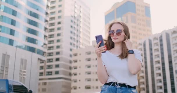 Joven hermosa mujer sonriendo con teléfono inteligente en la mano contra los rascacielos de verano — Vídeo de stock