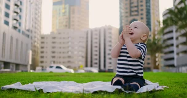 Happy small child sitting in grass with white daisies city background — Stockvideo