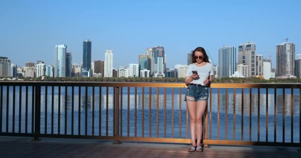 Chica en el fondo de la gran ciudad y la bahía sosteniendo un teléfono inteligente en gafas de sol — Vídeos de Stock