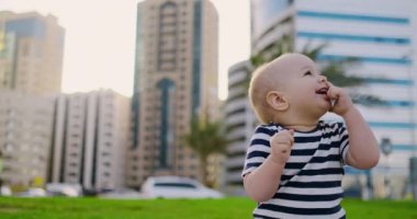 Erkek bebek yaz arka plan binalarda çim üzerinde oturan gülüyor