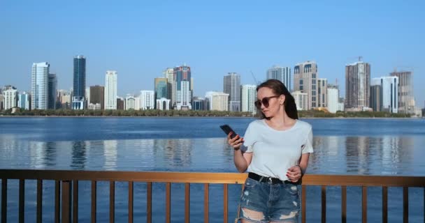 Junges schönes Mädchen, das am Wasser im Hintergrund der Stadt steht und ein Smartphone hält — Stockvideo