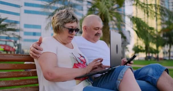 Heureux couple de personnes âgées assis sur un banc en été dans une ville moderne avec un ordinateur portable sur le fond des gratte-ciel — Video