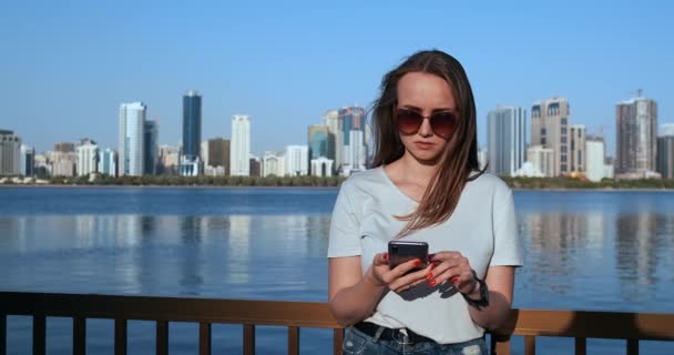 女孩在大城市和海湾的背景拿着智能手机在太阳镜 — 图库视频影像
