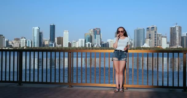 Fiatal gyönyörű lány áll a vízparton szemben a város beszél telefonon — Stock videók