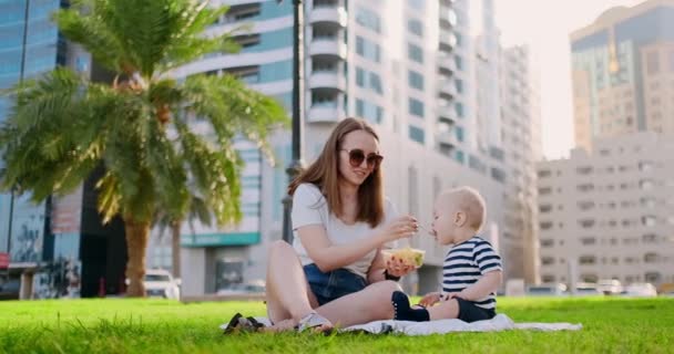 Молода мама з дитиною сидить на траві в парку їсть обід — стокове відео