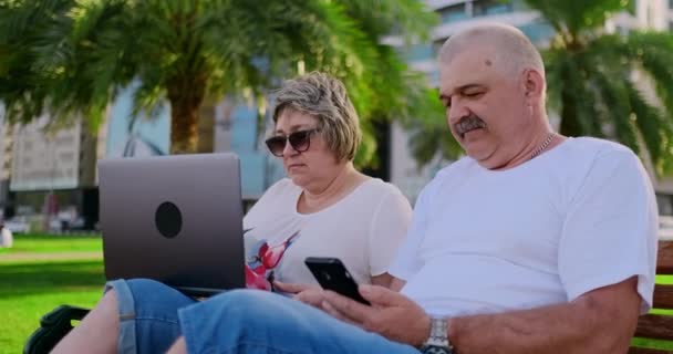 Heureux couple de personnes âgées assis sur un banc en été dans une ville moderne avec un ordinateur portable sur le fond des gratte-ciel — Video