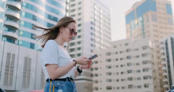 Joven hermosa mujer sonriendo con teléfono inteligente en la mano contra los rascacielos de verano — Vídeos de Stock