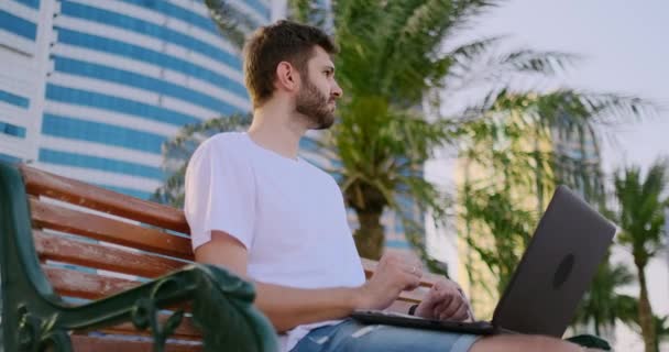 Un joven en el verano de palmeras sentado con un ordenador portátil y escribiendo en el teclado — Vídeo de stock