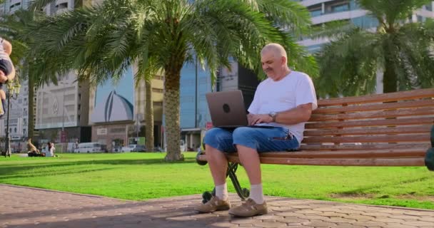 Hombre mayor sentado en un banco del parque entre las palmeras y mirando la pantalla del ordenador portátil — Vídeo de stock