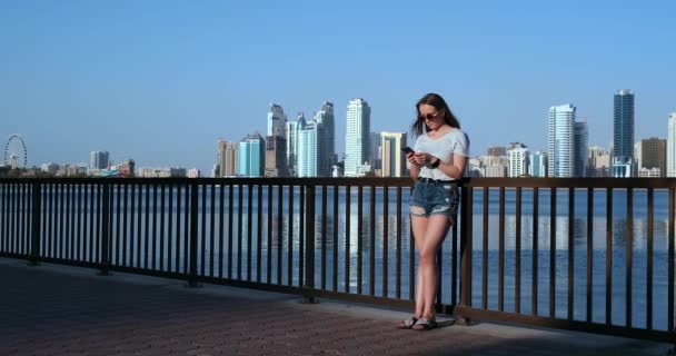 Mädchen auf dem Hintergrund der Großstadt und der Bucht mit einem Smartphone in der Sonnenbrille — Stockvideo