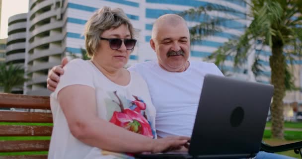 Feliz pareja de ancianos sentados en un banco en el verano en una ciudad moderna con un ordenador portátil entre las palmeras en el fondo de los rascacielos — Vídeo de stock