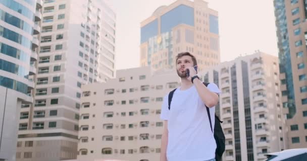 Egy fiatal férfi egy hátizsákot beszél a telefon a háttérben az épületek — Stock videók