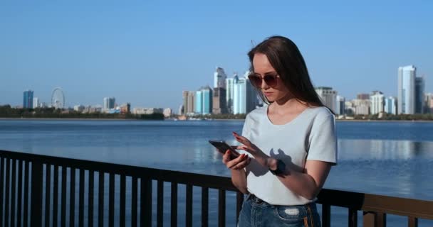 Mädchen auf dem Hintergrund der Großstadt und der Bucht mit einem Smartphone in der Sonnenbrille — Stockvideo